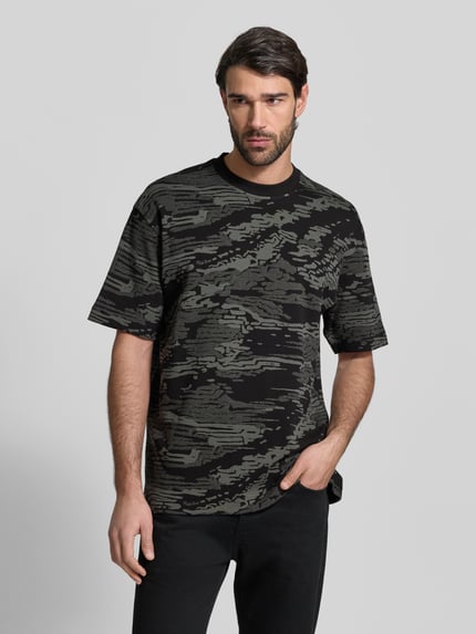 Męski model nosi Regular Fit T-Shirt aus reiner Baumwolle Modell 'TE_WILDNATURE' von BOSS Orange w abstrakcyjne, ciemnozielone i czarne wzory, krótki rękaw, okrągły dekolt, label patch, feinstrick, Ganzkörperansicht von vorne.