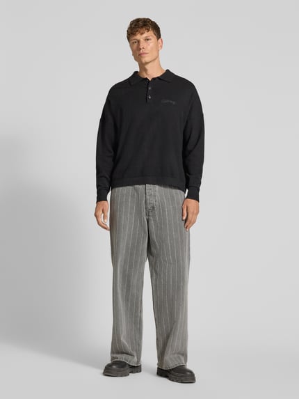 Mannenmodel draagt gebreide pullover met polokraag en knoopsluiting van RETERNITY, Materiaalmix met katoen, regular fit, labelstitching, ruglengte maat M 65 cm, mouwlengte maat M 48 cm, zwarte kleur, zachte textuur, rechte vorm, grijze broek met strepen, volledige lichaamsaanzicht van voren.