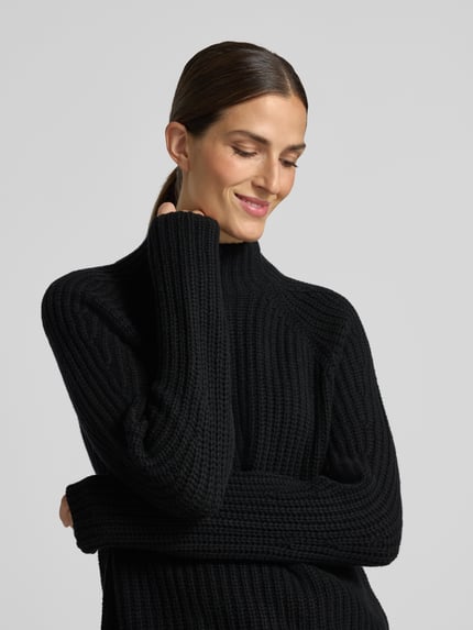 Frauenmodel trägt schwarzen, gerippten Strickpullover aus Kaschmir-Baumwoll-Mix Modell 'LONA' von 0039 Italy, Nahaufnahme Oberkörper. Regular Fit, Stehkragen, weiche Textur. Rückenlänge 60 cm, Ärmellänge 78 cm.
