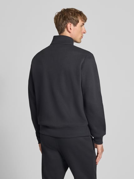 Męski model nosi Sweatanzug mit Label-Details od Champion, mieszanka bawełny, prosty krój, stójka, elastyczny pas, wpuszczane kieszenie, czarny kolor, widoczne logo, zdjęcie całej sylwetki z tyłu, Szerokość w rozmiarze M: 76,42 cm, Długość tyłu w rozmiarze M: 70 cm, Szerokość ramion w rozmiarze M: 51 cm, Długość rękawów w rozmiarze M: 69 cm, Długość wewnętrzna nogawki w rozmiarze M: 76 cm. Ganzkörperansicht Rückansicht