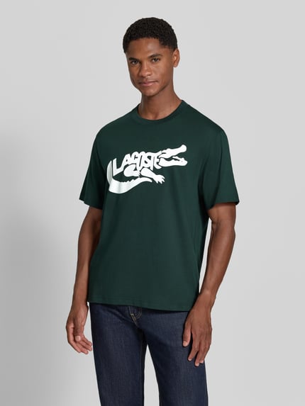 Männermodel trägt Regular Fit T-Shirt aus reiner Baumwolle von Lacoste in Dunkelgrün mit weißem Print, Nahaufnahme Oberkörper.