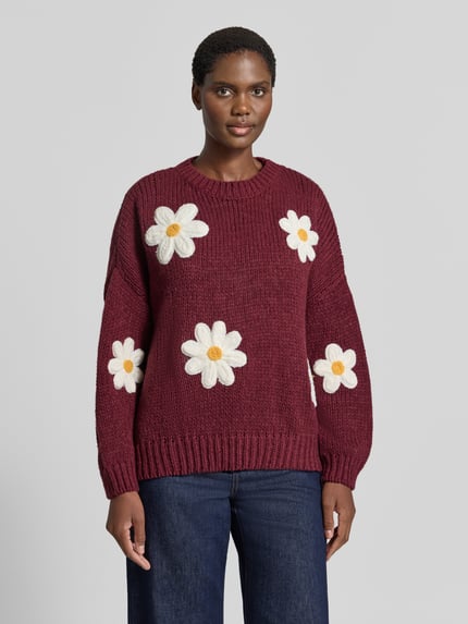 Frauenmodel trägt Oversized Strickpullover mit floralen Stitchings von Apricot in Dunkelrot mit weißen Blumen, grobe Stricktextur, Rundhalsausschnitt, breite Ärmel, gerippte Abschlüsse, Ganzkörperansicht von vorne.