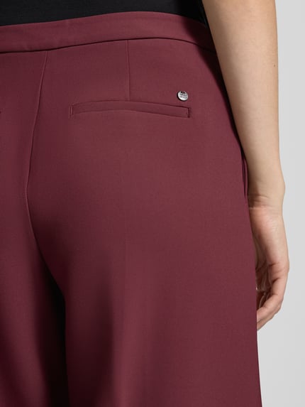 Frauenmodel trägt Wide Fit Hose mit Bügelfalten Modell 'Leya' von MOS MOSH in Bordeaux, glatte Textur, Detailaufnahme, Bundweite 62 cm, Innenbeinlänge 80 cm.