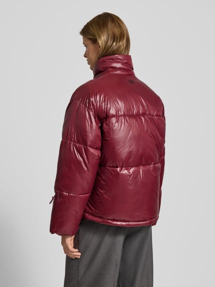 Kobieta modelka nosi kurtkę pikowaną ze stójką Model 'ALLIE' od Noisy May, krój relaxed fit, wpuszczane kieszenie, długość tyłu 66 cm, szerokość ramion 56 cm, klatki piersiowej 66 cm, długość rękawów 56 cm, gładka faktura, kolor bordowy, całkowity widok z tyłu.