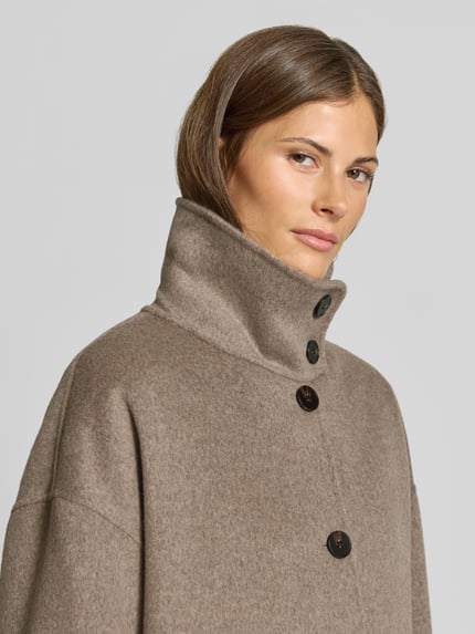 Frauenmodel trägt Wollmantel aus Woll-Mix von MYON in Beige mit hohem Stehkragen und Knopfleiste, Nahaufnahme Oberkörper. Lockere Passform, aufgesetzte Eingrifftaschen, Rückenlänge 115 cm, Schulterbreite 63 cm, Brustweite 65 cm, Ärmellänge 53 cm.
