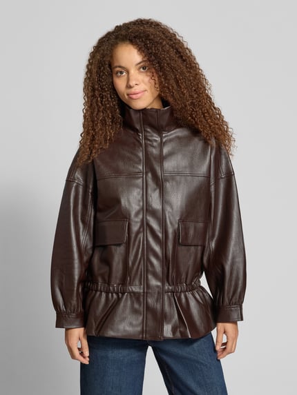 Vrouwenmodel draagt Jacke mit Stehkragen und Pattentaschen van Gina Tricot in leerlook, donkerbruin, getailleerd model, klepzakken, ritssluiting en drukknoopsluiting, oversneden schouders, ruglengte maat S 70 cm, schouderbreedte 70 cm, borstwijdte 60 cm, mouwlengte 46 cm, geheel vooraanzicht