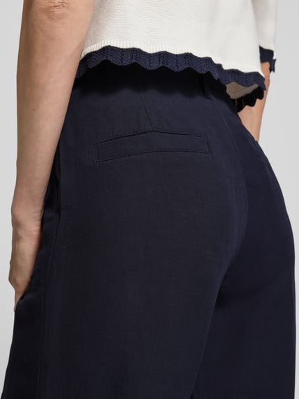 Kobieta nosi spodnie materiałowe Wide Fit Stoffhose aus Viskose-mix mit Leinen-Anteil od Marc Cain, mieszanka wiskozy i lnu, detal z logo, kieszenie z wypustkami, wpuszczane kieszenie, szerokość w rozmiarze 36: 37 cm, długość wewnętrzna nogawki w rozmiarze 36: 82 cm, ciemnogranatowy materiał, gładka tekstura, zbliżenie na tył spodni.