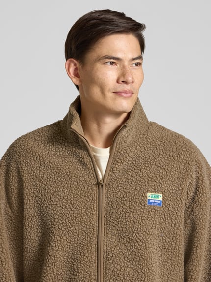Männermodel trägt Sweatjacke mit Logo-Patch Modell 'HOKTOWN' von AMERICAN VINTAGE, braune Fleece-Struktur, Nahaufnahme Oberkörper.