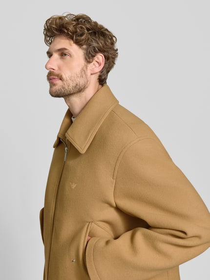 Männermodel trägt Jacke mit Logo-Stitching und Pattentaschen von Emporio Armani in Beige, strukturierte Oberfläche, Nahaufnahme Oberkörper. Gerader Schnitt, Kentkragen, Zweiwege-Reißverschluss, seitliche Pattentaschen, Rückenlänge 65 cm, Schulterbreite 52 cm, Brustweite 58 cm, Ärmellänge 70 cm.