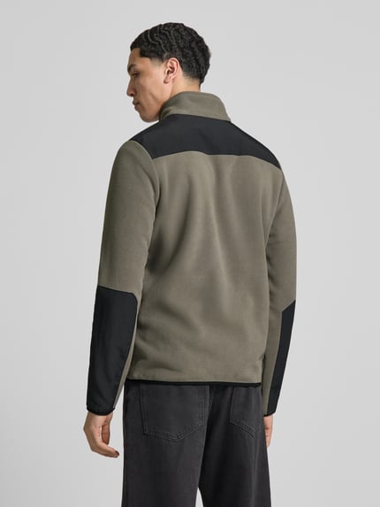 Męski model nosi kurtkę z polaru z stójką i kieszeniami zapinanymi na zamek błyskawiczny od HALO, krój regular fit, nadruk z logo, długość tyłu 71 cm, szerokość ramion 45 cm, szerokość w rozmiarze M 45 cm, długość rękawów 69 cm, kolor oliwkowy z czarnymi wstawkami, tekstura polarowa, widok całej sylwetki z tyłu.
