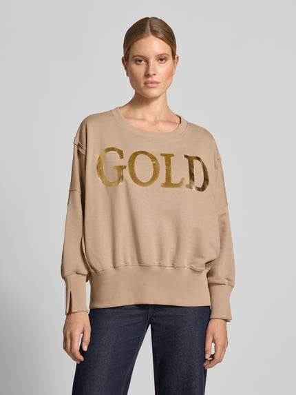 Vrouwenmodel draagt Oversized Sweatshirt uit puur katoen met statementprint van miss goodlife in beige met gouden letters, ronde hals, oversized fit, elastische boorden, voorkant bovenlichaam close-up.