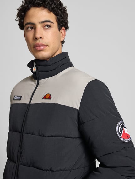 Männermodel trägt Steppjacke met rits Model 'NEBULA' van Ellesse in zwart-grijs met labeldetails, opstaande kraag, Nahaufnahme Oberkörper. Regular fit, zijzakken, ruglengte 77 cm, schouderbreedte 49 cm, borstwijdte 57 cm, mouwlengte 70 cm.