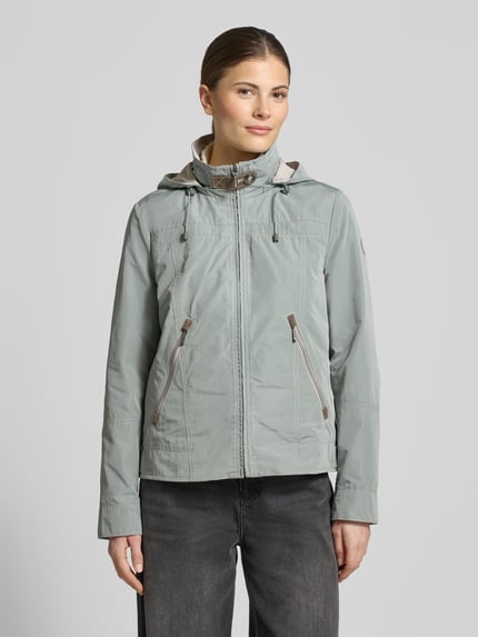 Modelka kobieta ma na sobie Jacke mit Kapuze und Reißverschlusstaschen od Gil Bret, krój regular fit, kolor jasnoszary, zamek, kaptur z troczkiem, kieszenie na suwak, stójka, długość tyłu w rozmiarze 36: 59 cm, szerokość ramion w rozmiarze 36: 40 cm, szerokość w klatce piersiowej w rozmiarze 36: 51 cm, długość rękawa w rozmiarze 36: 63 cm, widok całej sylwetki z przodu.
