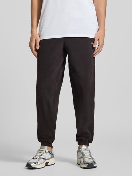 Männermodel trägt Relaxed Fit Sweathose mit Logo-Print Modell 'PUMATECH' von PUMA, schwarze Farbe, lockere Passform, Herren Sweathose, Nahaufnahme Unterkörper, Vorderansicht.
