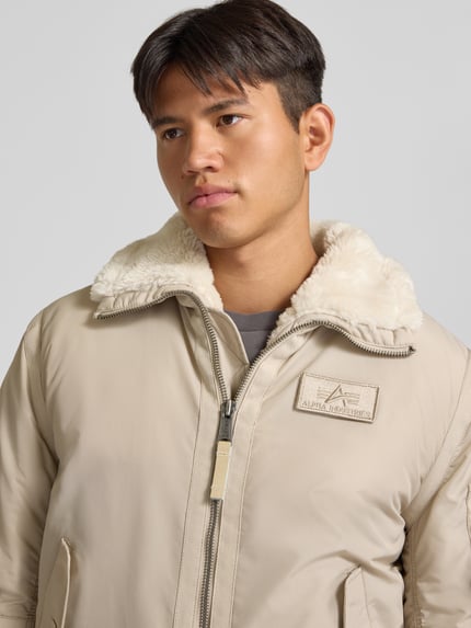 Männermodel trägt Bomberjacke mit Label-Patch und Kentkragen von Alpha Industries in Beige, sichtbarer Teddyfellkragen, Nahaufnahme Oberkörper. Regular Fit, Reißverschluss, Brustweite 56 cm, KI-modifiziertes Bild.