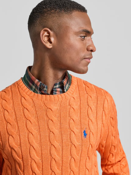 Męski model nosi Strickpullover mit Label-Stitching od Polo Ralph Lauren, bawełna, regular fit, pomarańczowy, detal na piersi, zbliżenie na górę ciała.