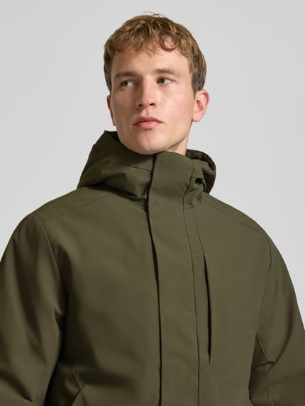 Męski model nosi kurtkę z kapturem, zakrytym zamkiem, model 'KEEN' od JACK & JONES, krój regular fit, z paskiem na rękawie, kolor oliwkowy, z teksturą gładką, szerokość w rozmiarze M 60 cm, długość tyłu 72 cm, szerokość ramion 49 cm, długość rękawa 68 cm, z przodu zbliżenie górnej części ciała.