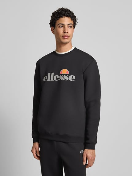 Männermodel draagt zwart Sweatshirt met label-print model 'CORVARO' van Ellesse, ronde hals, ribboorden, losse pasvorm, materiaal-mix, voorkant bovenlichaam close-up.