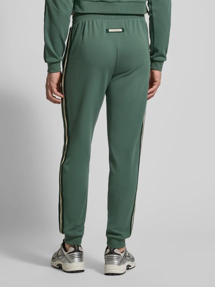 Męski model nosi spodnie dresowe z mieszanki bawełny i elastanu Sweatpants aus Baumwoll-Mix od EA7 Emporio Armani, luźny krój, elastyczny pas z tunelem, elastyczne zakończenia nogawek, wpuszczane kieszenie, długość wewnętrzna nogawki w rozmiarze M: 78 cm, zielony kolor, gładka tekstura, widok całej sylwetki z tyłu.