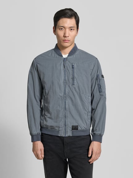 Mannenmodel draagt Blouson met Label-Patches Modell 'IBE' van khujo in grijs, rechte fit, korte opstaande kraag, rits, borstzak met rits, mouwzak, zijzakken, gladde textuur. Volledig vooraanzicht borst tot heup.