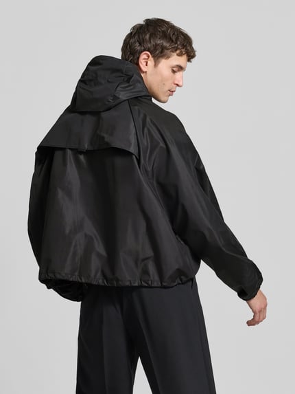 Männermodel trägt kurtka przeciwdeszczowa z kieszeniami z patką od VETEMENTS, czarna, luźny krój, Ganzkörperansicht Rückansicht, Długość tyłu w rozmiarze L: 66 cm, Szerokość ramion w rozmiarze L: 42 cm, Długość rękawów w rozmiarze L: 80 cm