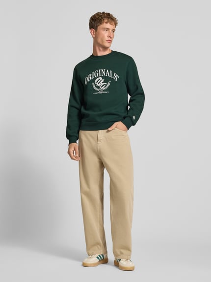 Männermodel trägt Sweatshirt mit Label-Print Modell 'FREDERIKSBERG' von JACK & JONES, dunkles Grün mit beigem Print, Rundhals, Baumwoll-Mix, Regular Fit, beigefarbene Hose, Ganzkörperansicht von vorne.