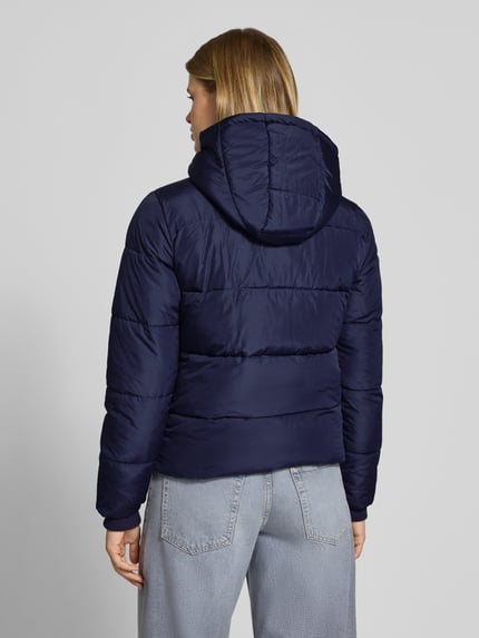 Frauenmodel trägt Steppjacke mit Kapuze von PIECES in Dunkelblau, gesteppte Struktur, kombiniert mit hellblauer Jeans, Ganzkörperansicht Rückansicht.
