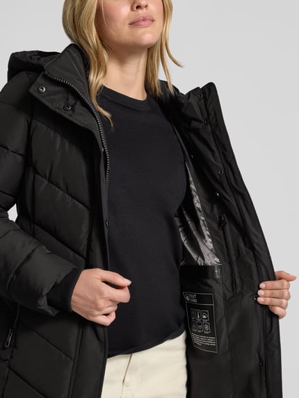 Frauenmodel trägt Jacke mit abnehmbarer Kapuze von NAUTGÅRD in Schwarz, gesteppt, glatte Textur, Nahaufnahme Oberkörper.