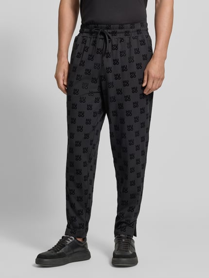 Männermodel trägt Regular Fit Sweathose aus Baumwoll-Mix von HUGO in Schwarz mit Allover-Logomuster und elastischem Bund, Nahaufnahme Unterkörper. Herren Sweathose von HUGO.