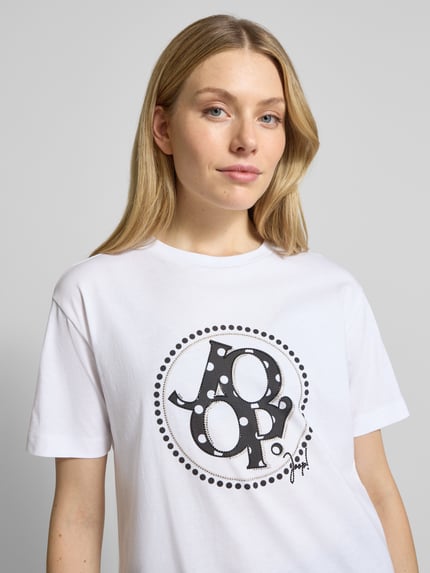 Frauenmodel trägt T-Shirt mit Ziersteinbesatz Modell 'Tammi' von JOOP!, weiß, Rundhalsausschnitt, Regular Fit, Nahaufnahme Oberkörper.
