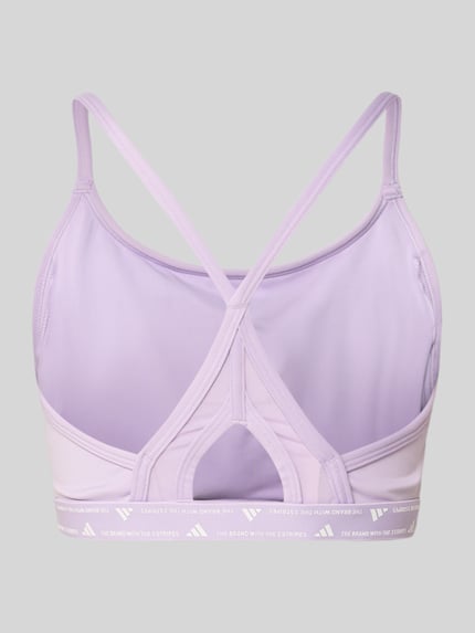 Vrouwenmodel draagt Bh met Labelprint van adidas Sportswear in lila, materiaalmix met stretch, nauwsluitend model, elastische band. Ganzkörperansicht Rückansicht.
