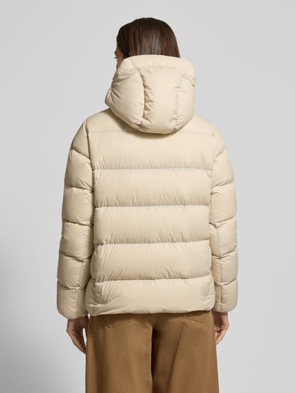 Frauenmodel trägt beige Steppjacke mit Kapuze, Label-Stitching und Reißverschluss, Modell 'FROST DOWN' von PEAK PERFORMANCE, Regular Fit, Rückenlänge 64 cm, Schulterbreite 41 cm, Brustweite 56 cm, Ärmellänge 64 cm, Ganzkörperansicht Rückansicht.