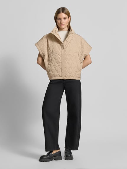 Vrouwenmodel draagt Cape met Steppnähten und Eingrifftaschen van Copenhagen Muse in beige met doorgestikte naden en steekzakken, relaxed fit, hooggesloten kraag, knoopsluiting, ruglengte 66 cm, schouderbreedte 42 cm, borstwijdte 25 cm, Ganzkörperansicht van voren.