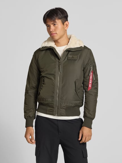 Männermodel trägt Bomberjacke mit Label-Patch und Kentkragen von Alpha Industries in Olivgrün, Teddyfellkragen, Ganzkörperansicht von vorne. Regular Fit, Reißverschluss, gerippte Abschlüsse, Reißverschlusstasche und Eingrifftaschen, Brustweite 56 cm bei Größe M.