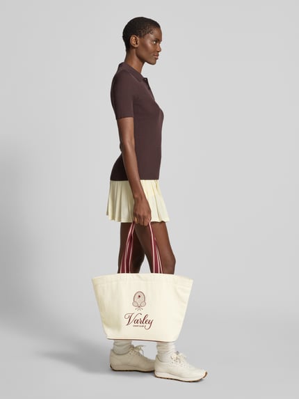 Frauenmodel trägt Shopper aus reiner Baumwolle Modell 'Robinson' von VARLEY in Creme mit Motiv-Stickereien, seitliche Ganzkörperansicht. Hauptfach mit Karabinerverschluss, Reißverschlussfach innen, Lange Tragehenkel, Label-Details.