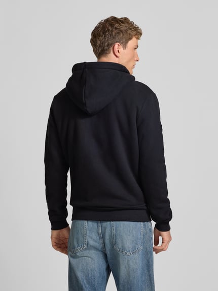 Männermodel trägt Regular Fit Sweatjacke mit geteilter Känguru-Tasche Modell 'Borg' von Superdry in Schwarz, jeansblaue Hose, Rückenlänge 71 cm, Ärmellänge 72 cm, Ganzkörperansicht Rückansicht.