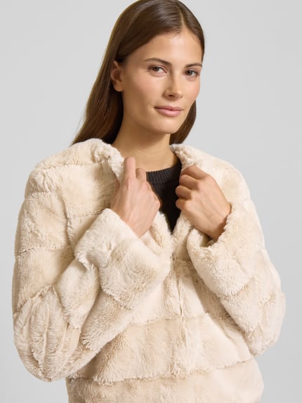 Kobiece modelka nosi Kurtka aus Kunstfell mit Rundhalsausschnitt od More & More, sztuczne futro, krój regular fit, okrągły dekolt, pikowanie, długość tyłu 53 cm, szerokość ramion 40 cm, szerokość klatki piersiowej 47 cm, długość rękawów 61 cm, kremowy kolor, tekstura puszysta, zbliżenie na górną część ciała
