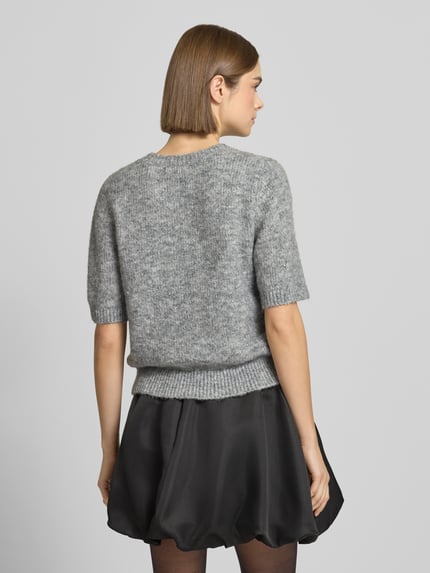 Frauenmodel trägt Strickpullover mit 1/2-Arm und Rundhalsausschnitt von Gina Tricot im grauen Material-Mix mit Lanawoll-Anteil, Regular-Fit, Ziersteinbesatz, kombiniert mit schwarzem Rock, Rückenlänge bei Größe S 53 cm, Ärmellänge 28 cm, Ganzkörperansicht Rückansicht.