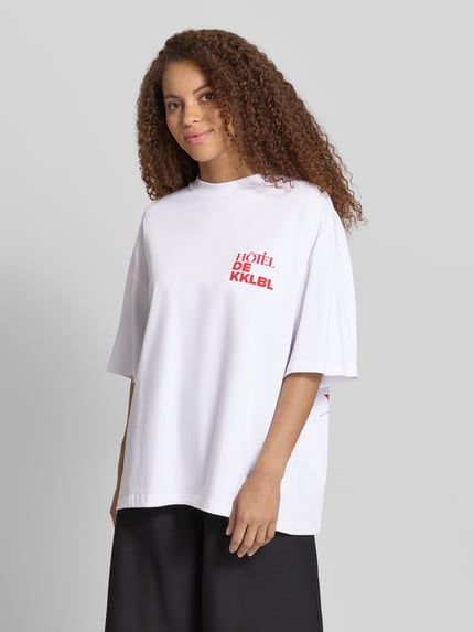 Frauenmodel trägt Karo Kauer Damen T-Shirt mit rückseitigem Print Modell 'Vacation' aus Reine Baumwolle, Oversized-Fit, Rundhalsausschnitt, Vorderseite mit rotem Statement-Print, glatte Textur, Weiß mit roten Akzenten, Ganzkörperansicht von vorne.