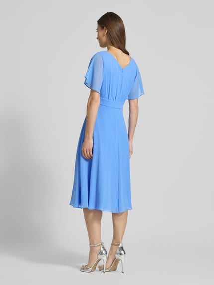 Vrouwenmodel draagt Knielanges Kleid mit Rundhalsausschnitt van Vera Mont, semi-transparant design, getailleerde fit, korte mouwen, blinde ritssluiting, ondoorzichtige onderjurk, ruglengte 102 cm, borstwijdte 37 cm, mouwlengte 27 cm, blauwe kleur, vloeiende stof, glanzende zilveren sandalen, volledige lichaamsweergave achteraan.