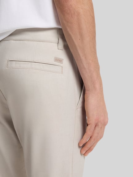 Männermodel trägt Slim Fit Chino aus Baumwoll-Mix mit Leinen und Viskose-Anteil von BOSS Orange in heller Farbe, Label-Patch, Nahaufnahme Rückansicht. Denim-Optik, Passform Slim Fit, Mid Waist, seitliche Eingrifftaschen, Gesäßtaschen, Materialverarbeitung Twill.