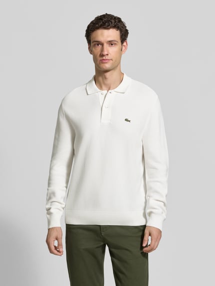 Męski model nosi Regular Fit Poloshirt aus reiner Baumwolle von Lacoste, biała koszulka polo, zielone spodnie, gładka faktura, Ganzkörperansicht von vorne.
