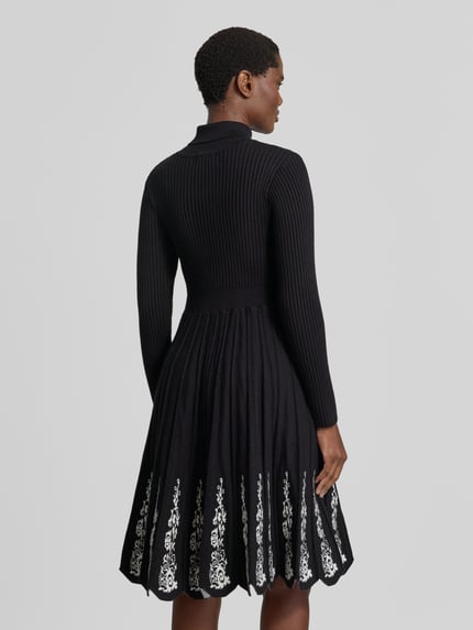 Frauenmodel trägt Knielanges Strickkleid aus Viskose-Mix von Apricot, schwarzer Stoff, weiße florale Stickereien, Ganzkörperansicht Rückansicht.