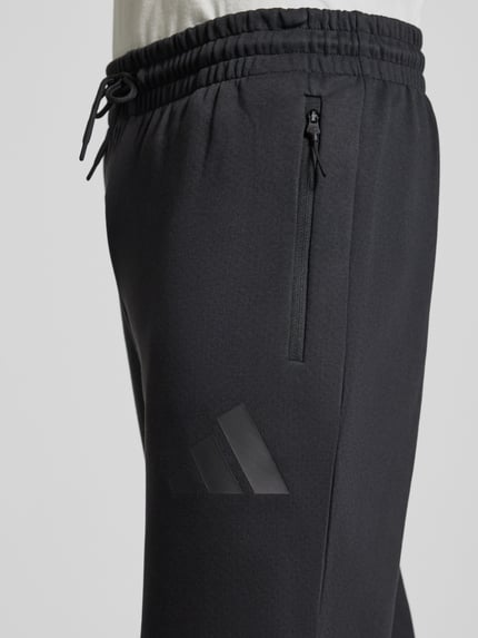 Männermodel trägt Sweatpants mit Logo und elastischem Bund von adidas Sportswear, Materiaalmix met katoen, regular fit, logoprint, elastische band, tunnelkoord, binnenbeenlengte maat M 75 cm. Detailaufnahme rechter heup.