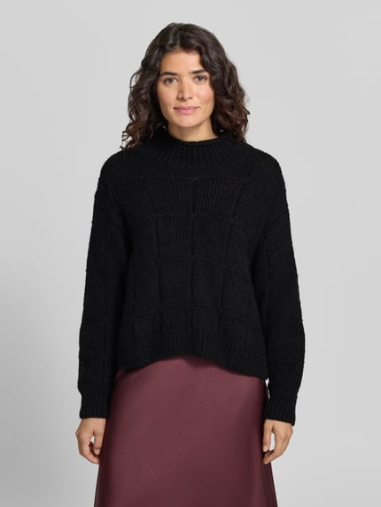 Kobiece modelka nosi sweter wełniany z strukturą dzianiny 'Pullover aus Wolle mit Strukturstrick' od MARC CAIN, jednokolorowy, imitacja dzianiny, pudełkowy krój, skrócona długość, szwy działowe, opuszczone ramiona, kołnierz mock neck, wysoka stójka, długie rękawy z prążkowanymi wykończeniami, prążkowane wykończenie dołu, widok całej sylwetki z przodu.