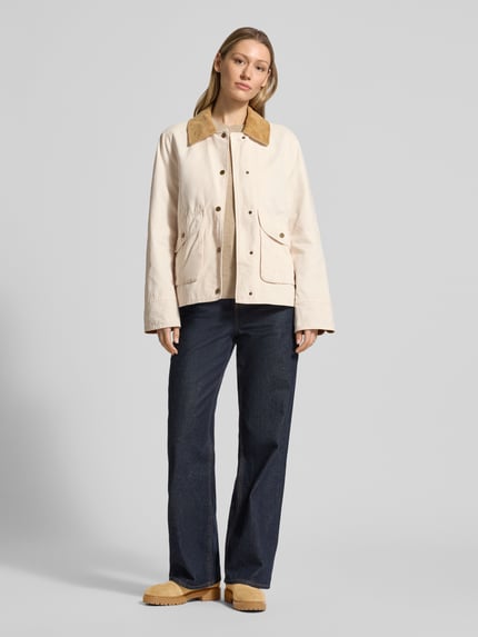 Vrouwenmodel draagt Jacke mit Umlegekragen und aufgesetzten Pattentaschen van WHITE LABEL in recht model met platte kraag, drukknoopsluiting, opgestikte klepzakken, ruglengte maat 36: 67 cm, schouderbreedte: 44 cm, borstwijdte: 53 cm, mouwlengte: 63 cm, lichte crèmekleuren, gladde textuur, donkere broek, bruine schoenen. Volledig frontaal totaalbeeld.