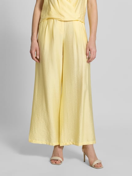 Frauenmodel trägt Wide Leg Pants aus Viskose-Mix mit Eingrifftaschen von GUIDO MARIA KRETSCHMER WOMAN, gelb, ausgestellter Schnitt, Nahaufnahme Oberkörper bis Fuß, Kontext: Modell 'Finja' mit Knopf-, Reißverschluss und französischen Eingrifftaschen.