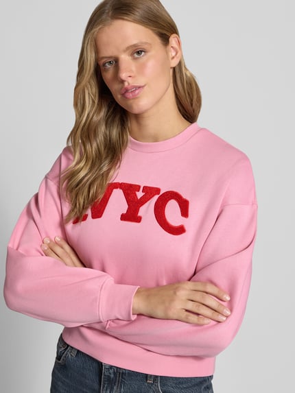 Frauenmodel trägt Relaxed Fit Sweatshirt aus Baumwoll-Mix Modell 'Tatum' von JJXX, pink mit rotem Statement-Patch, Rundhalsausschnitt, Rippbündchen, langärmlig, überschnittene Schultern, Nahaufnahme Oberkörper.