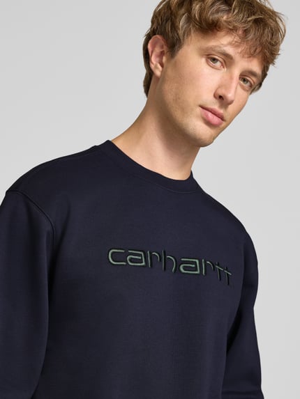 Männermodel trägt Regular Fit Sweatshirt aus Baumwoll-Mix mit Logo-Stickerei von Carhartt Work In Progress in Dunkelblau, Nahaufnahme Oberkörper. Baumwoll-Mix, gerippte Abschlüsse, langärmliges Design, überschnittene Schultern, Rundhalsausschnitt, gerippte Blende, Logo-Stickerei, Rückenlänge 70 cm, Ärmellänge 66 cm.