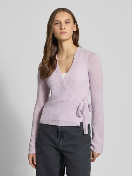 Frauenmodel trägt Strick-Cardigan in Wickeloptik von Review in Flieder, V-Ausschnitt, lange Ärmel, Schleifenverschluss, Ganzkörperansicht von vorne.